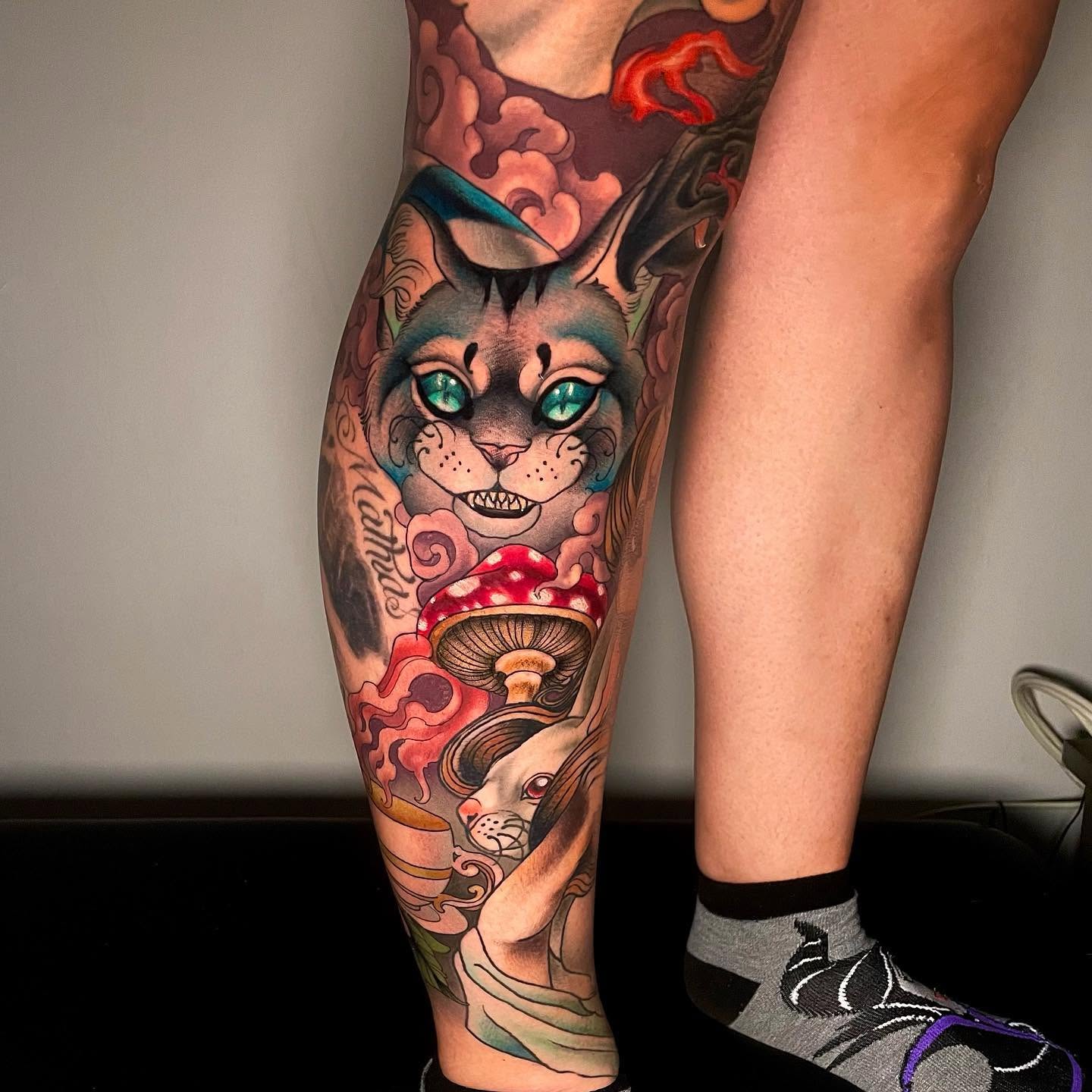 Close crop of a calf tattoo: a Cheshire cat with turquoise eyes above a red-capped mushroom and a white rabbit