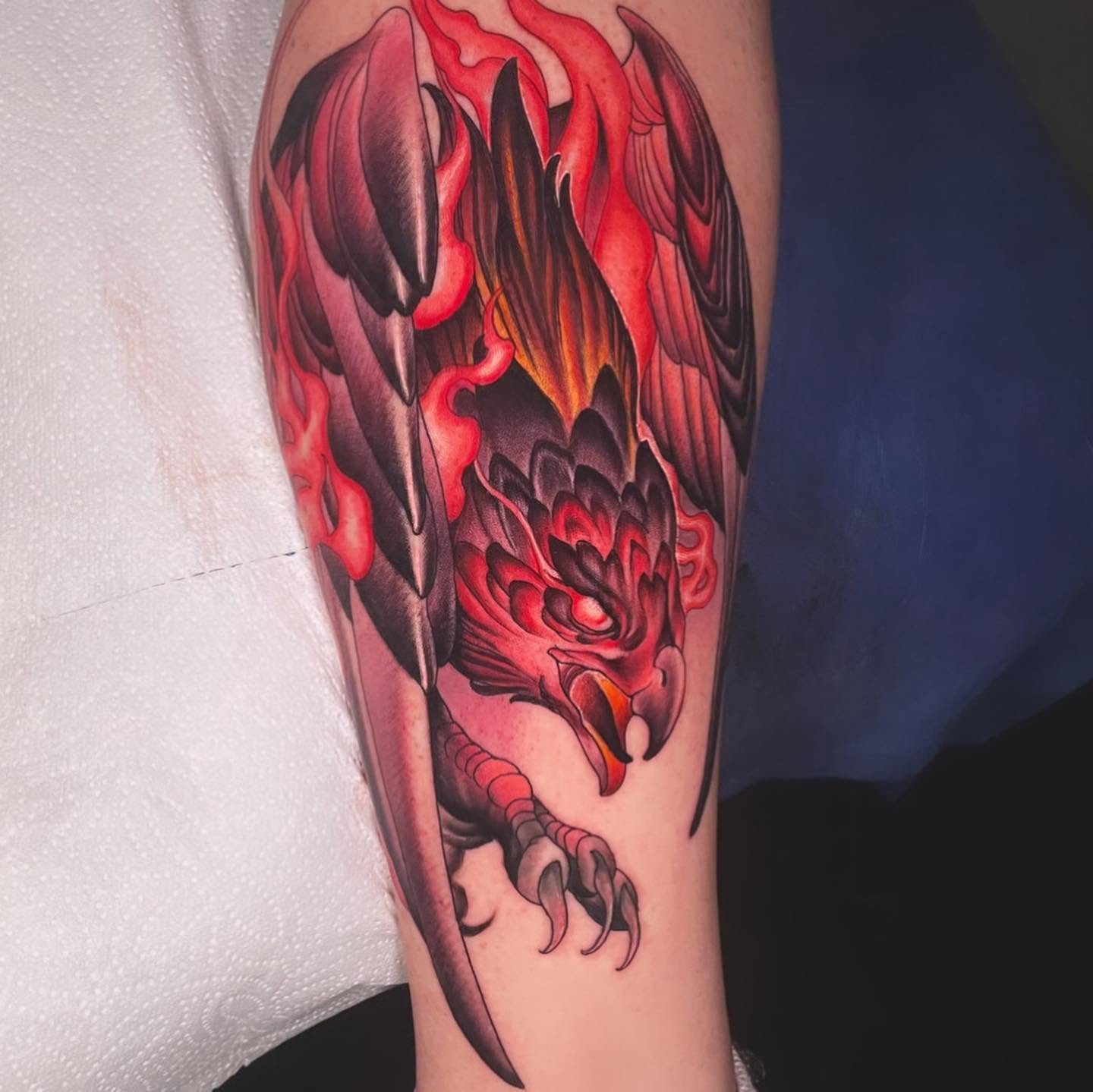 Arm tattoo of a crimson hawk diving with open talons through red flame feathers