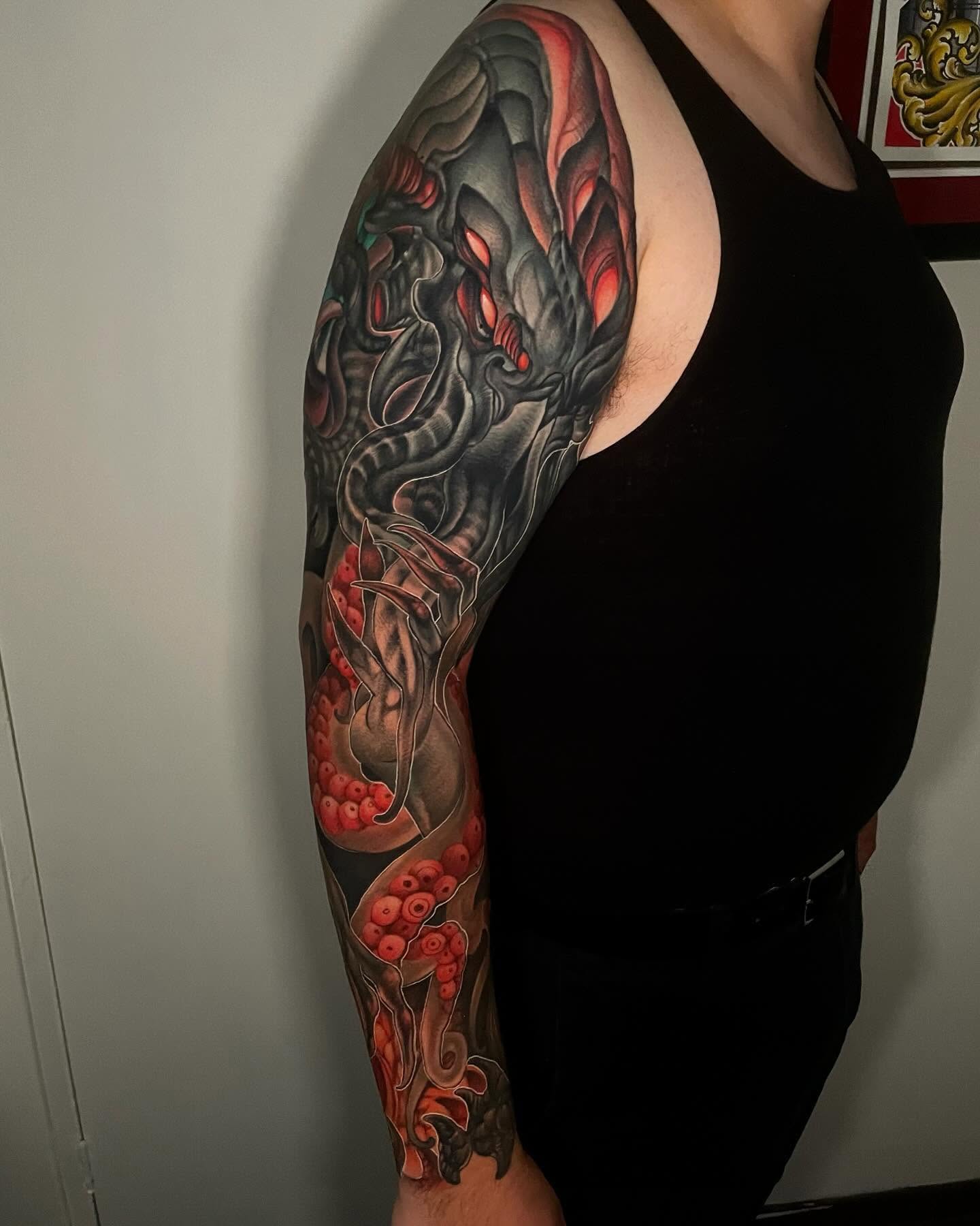 Full-sleeve tattoo of a black biomechanical creature with glowing red eyes and coral-red tentacles
