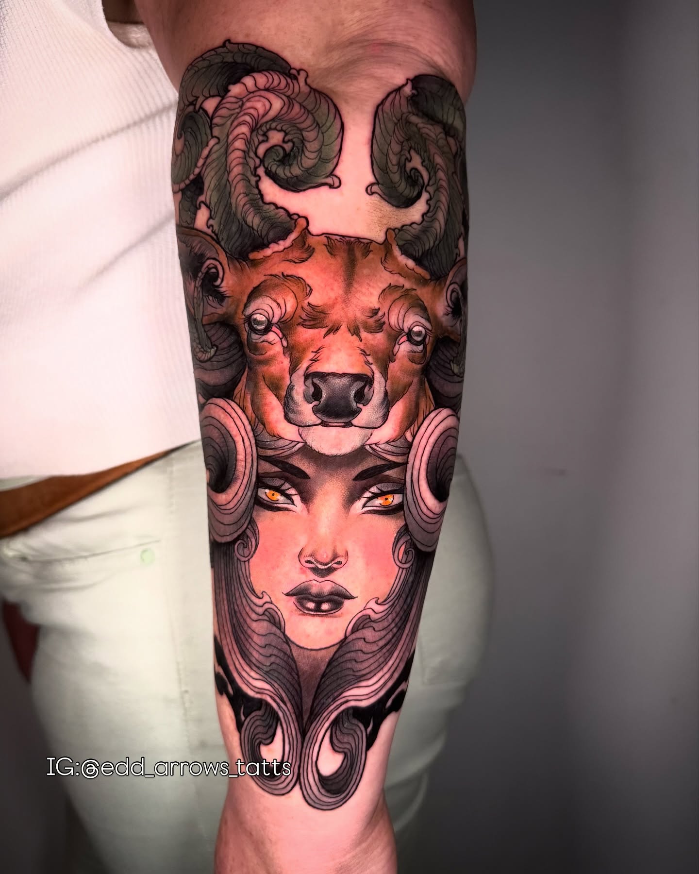 Forearm tattoo of a ram's head above a woman's face with amber eyes, wrapped in smoky horns
