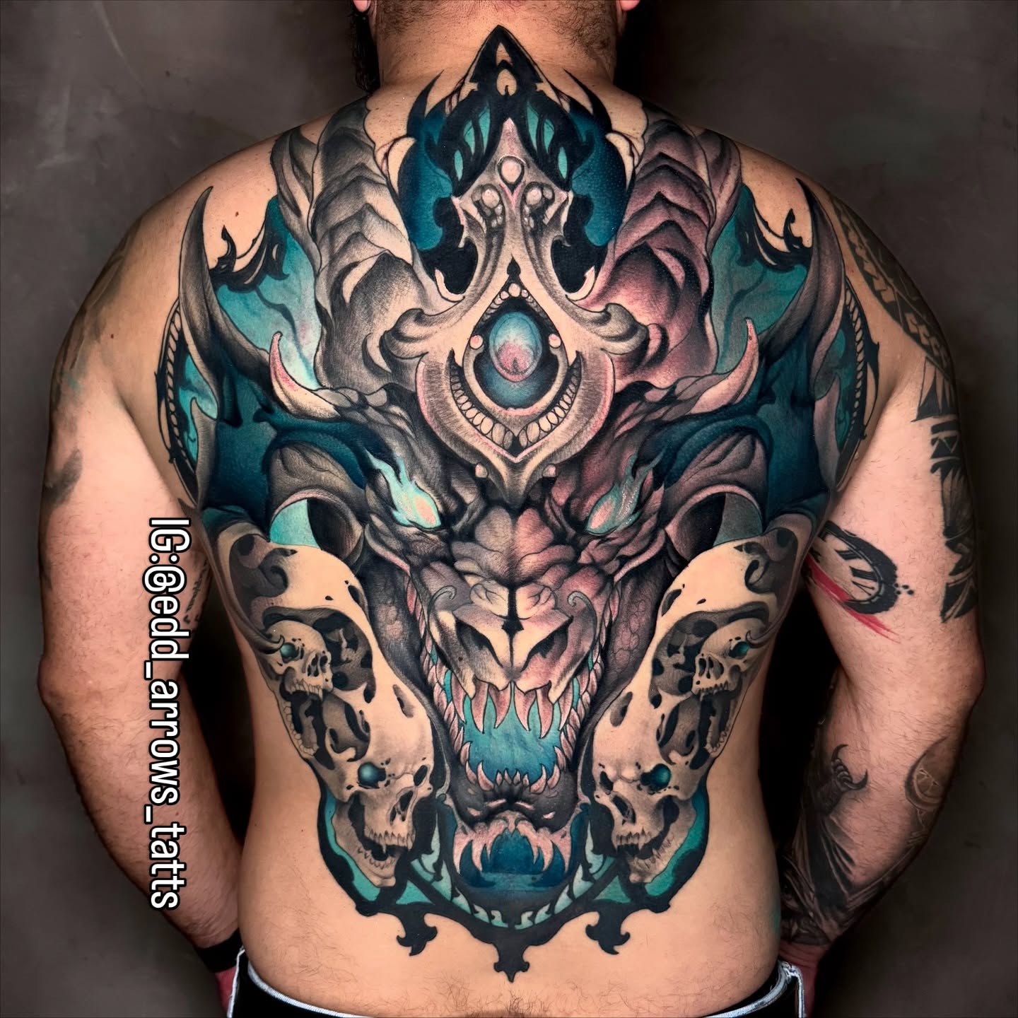 Full-back tattoo of a horned dragon with glowing teal accents, flanked by two animal skulls, in neo-traditional color