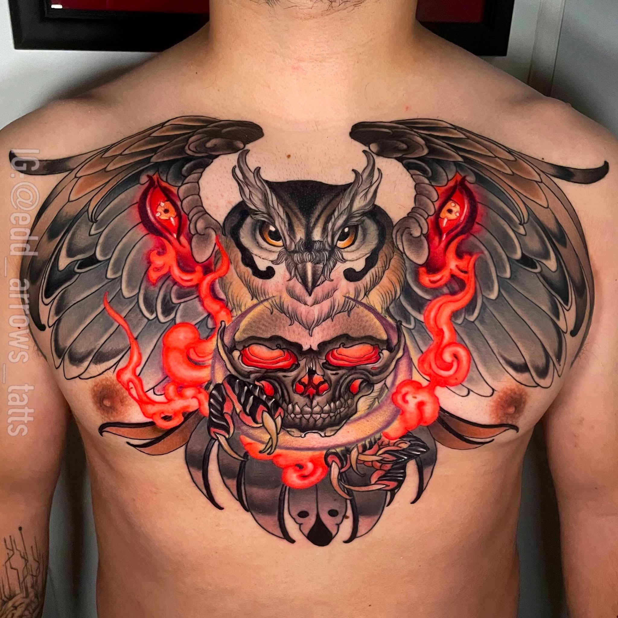 Chest tattoo of a horned owl with spread wings holding a skull, red flames curling around it, in neo-traditional color